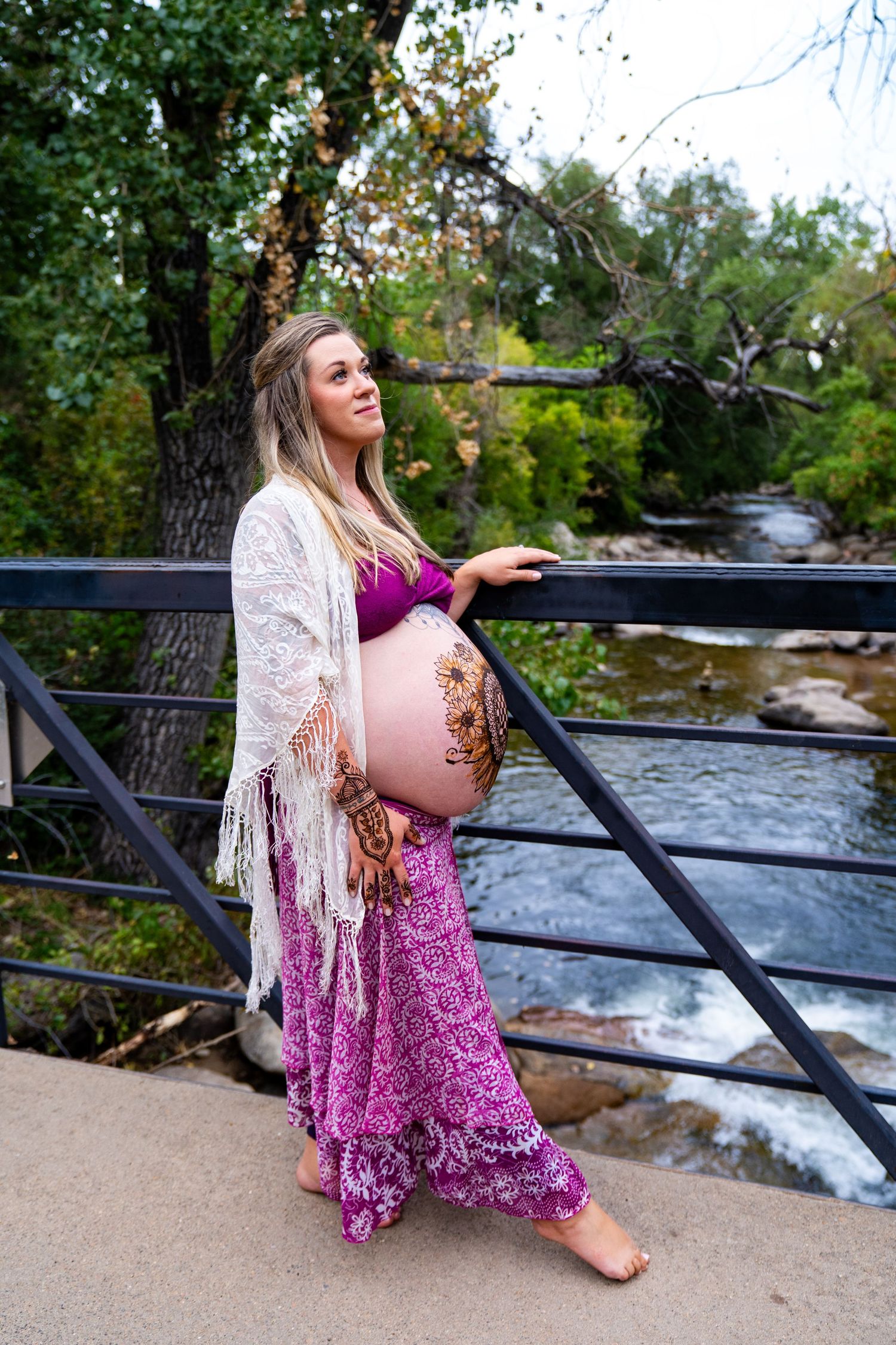 Expectant mother in flowing pink dress and lace shawl poses by river during autumn maternity photoshoot.
