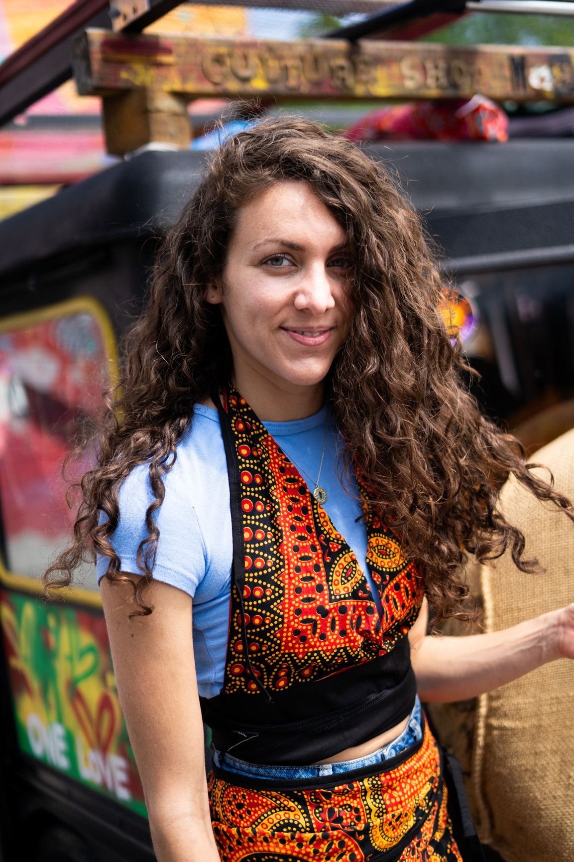 A photo of a woman with brown wavy hair looking at the camera and smiling and wearing a patterned  dress.