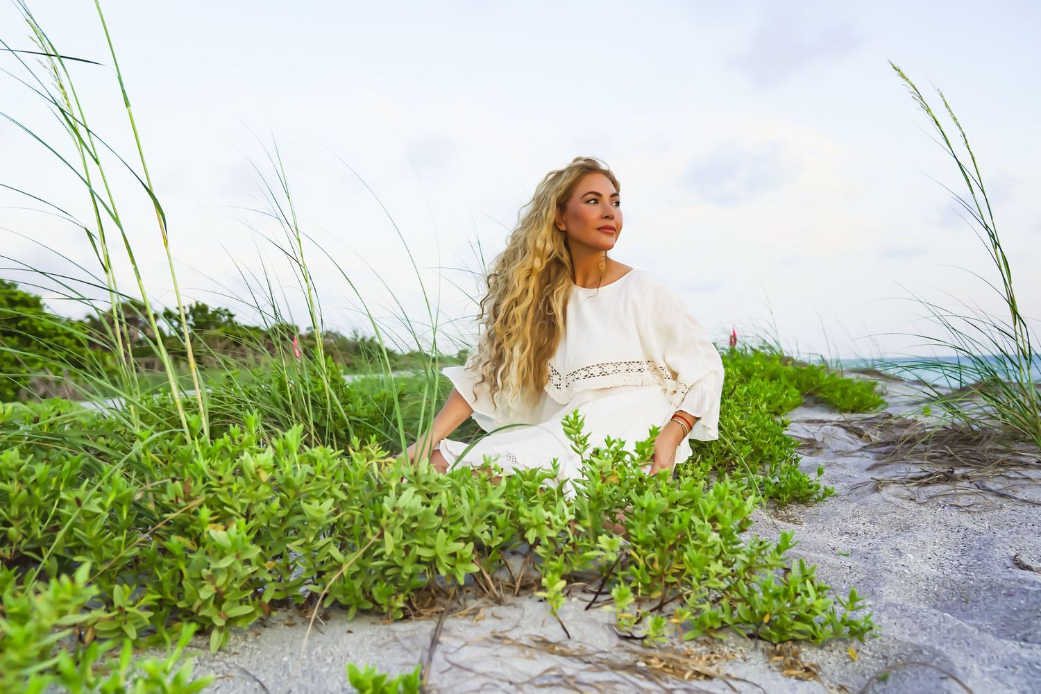 A photo of a woman with blonde flowy hair on the beach wearing a white outfit sitting around lots of greenery.