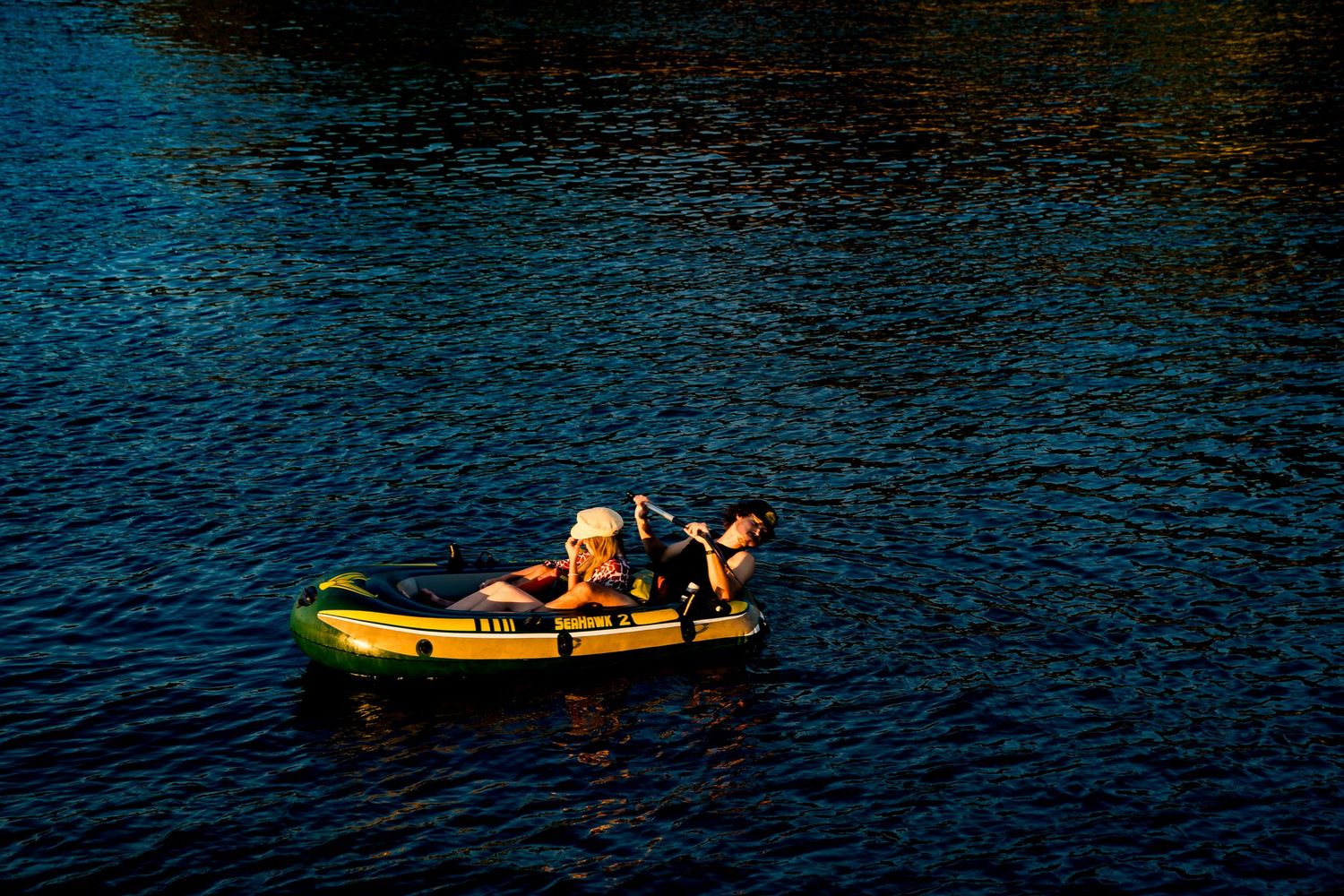 Two people relaxing in a yellow inflatable kayak on dark blue water, bathed in warm golden sunlight.