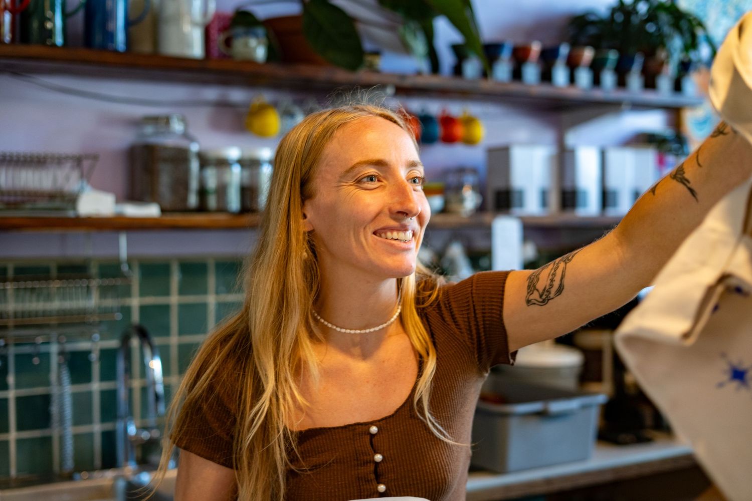 Smiling woman with long blonde hair working behind a cafe counter, wearing a brown top and pearl necklace.