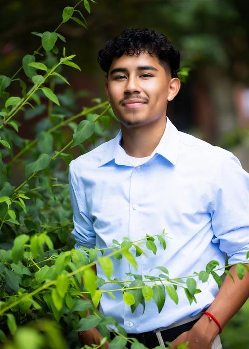 Young man in light blue shirt smiling outdoors surrounded by lush green foliage in a natural garden setting.