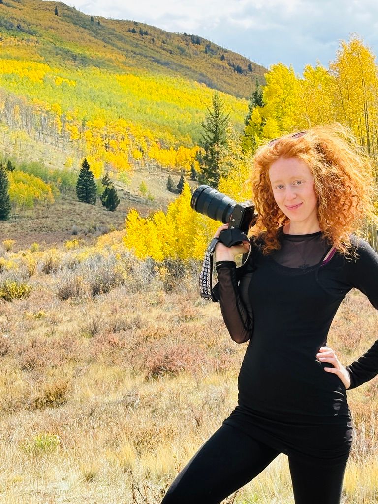 Red-haired woman holding a camera in black outfit, posing in front of vibrant fall foliage in Colorado mountains.