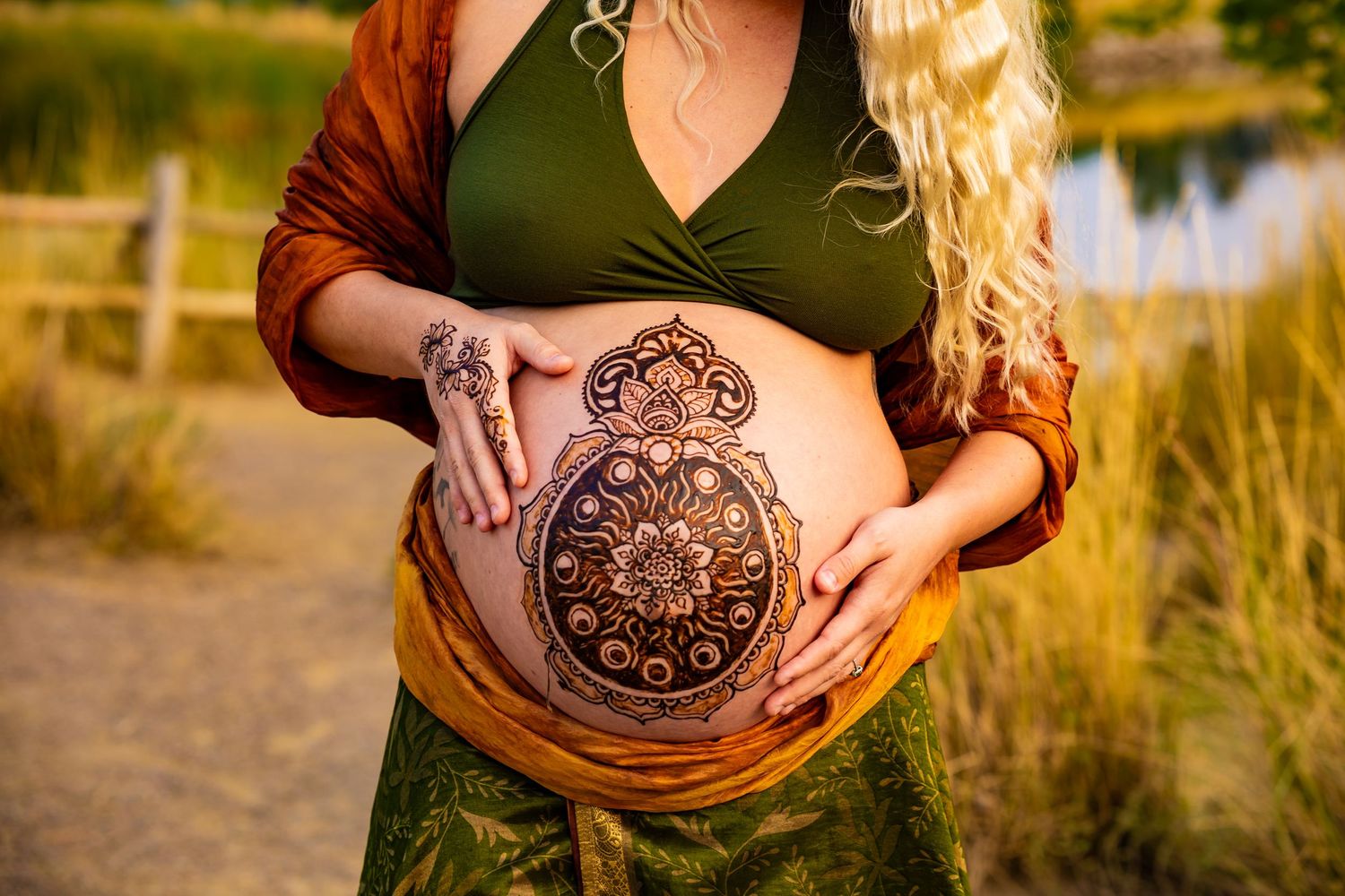 A pregnant belly adorned with intricate henna art designs captured in warm golden sunlight by a lake.