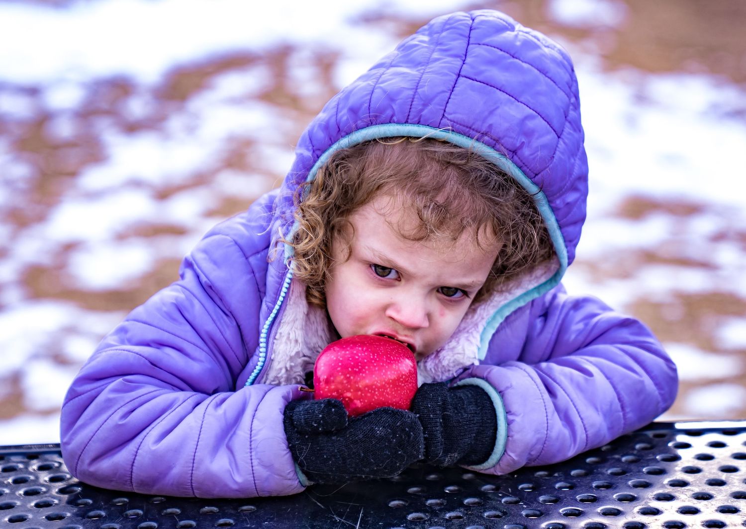 A child in a purple winter coat rests on a table while holding a red snow cone in winter.