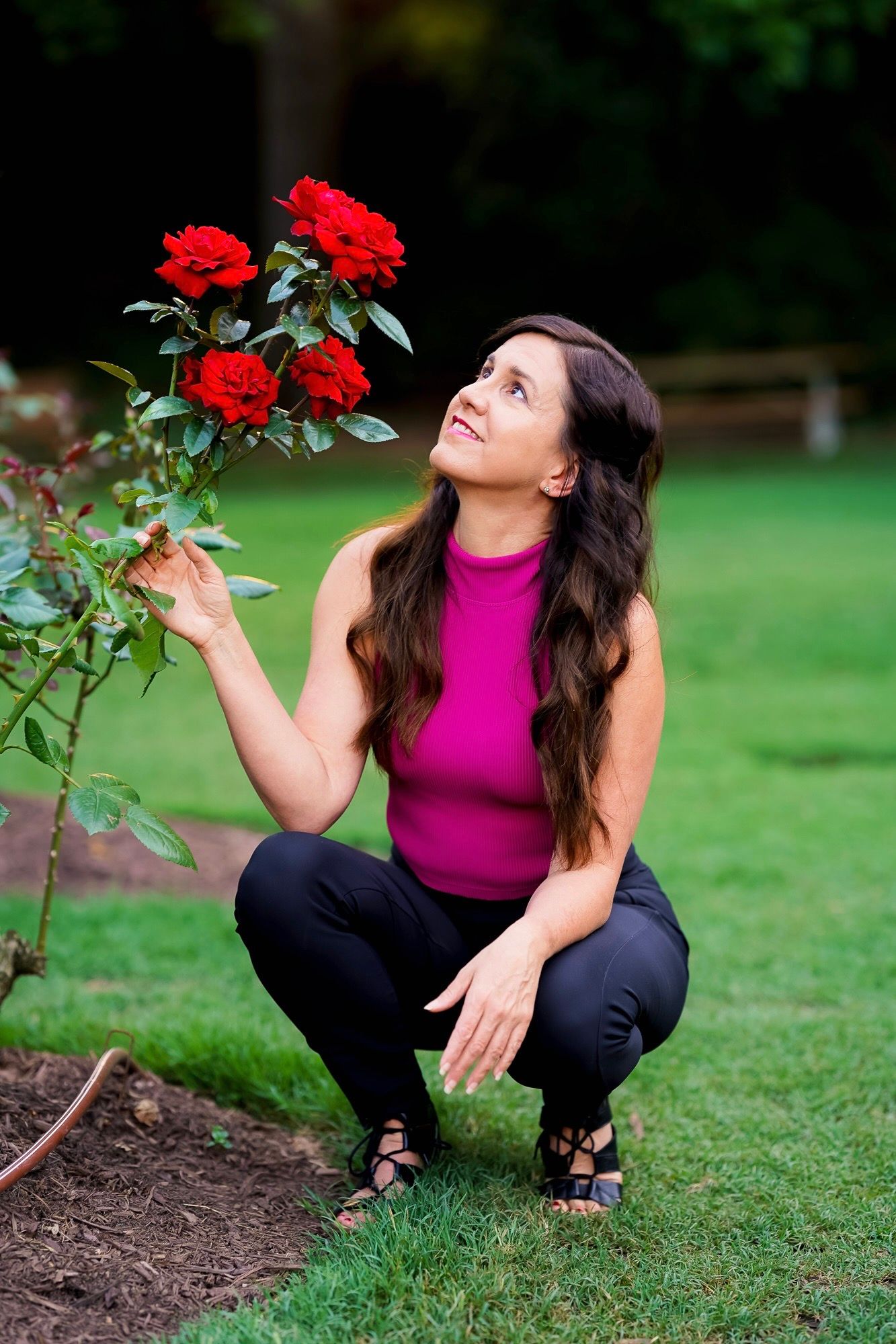A person in a bright pink sleeveless top crouches near vibrant red roses in a garden setting.