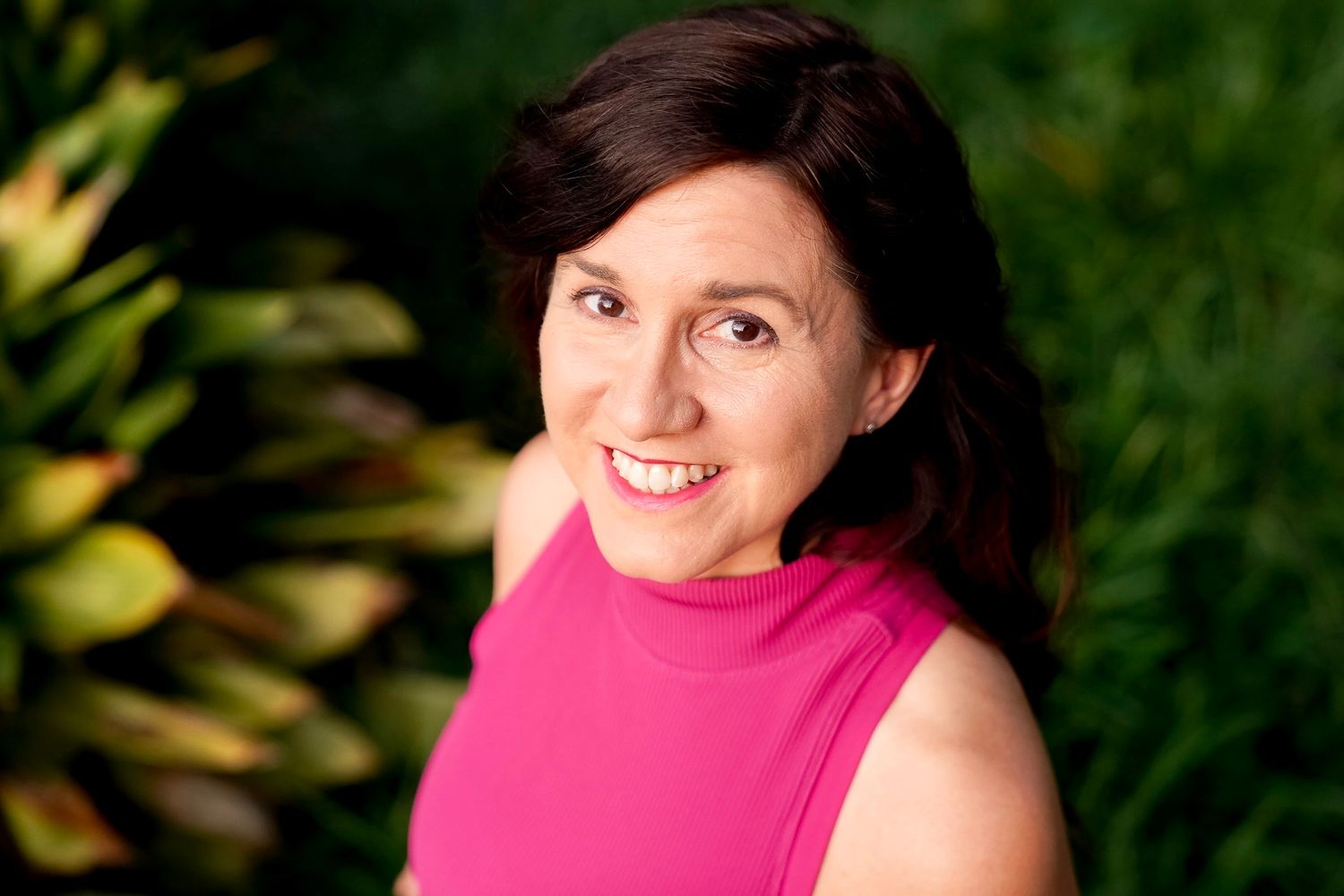 A person in a bright pink top smiles warmly while posing outdoors against a natural green background.