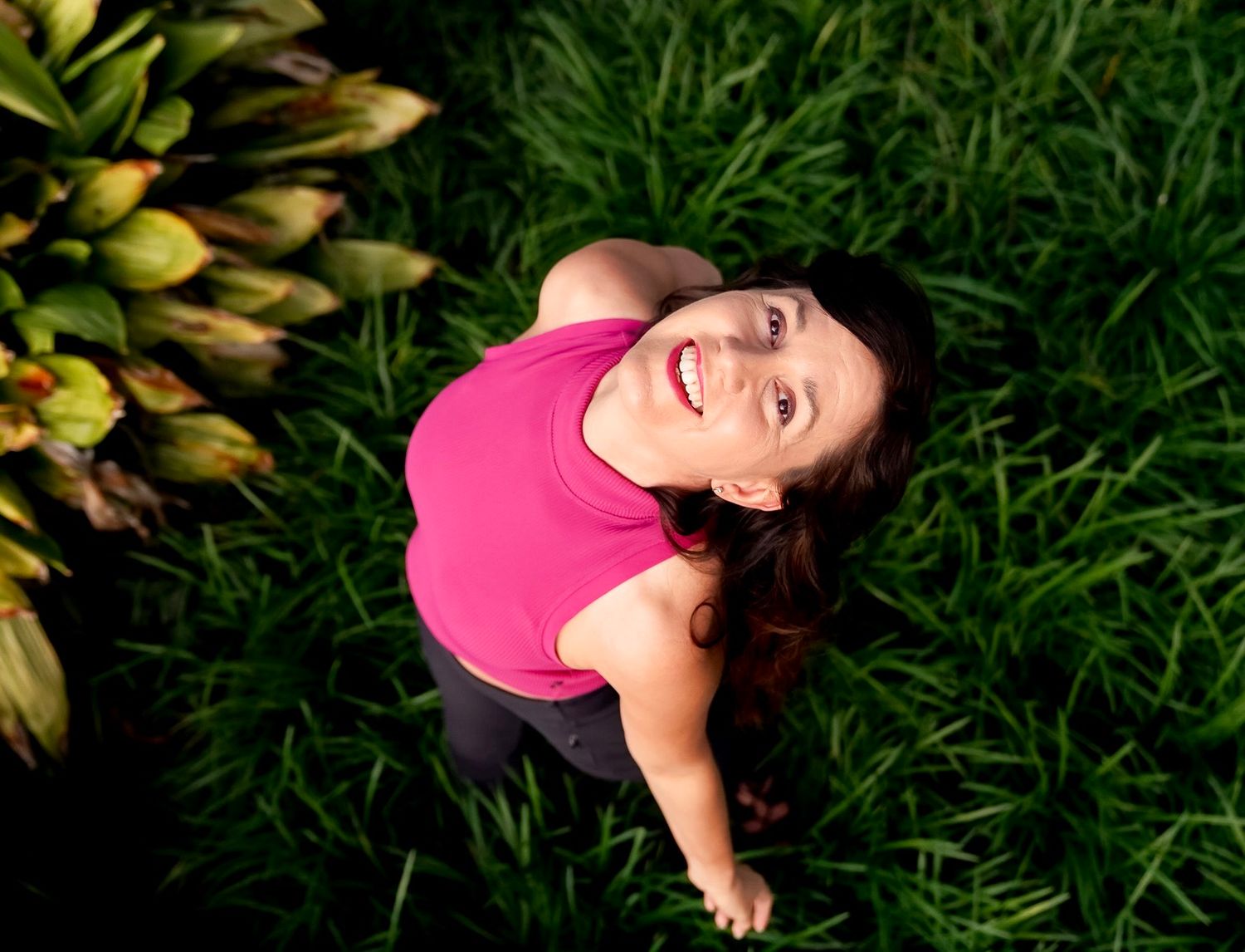 Person lying in green grass wearing pink shirt and dark pants enjoying a sunny day outdoors.