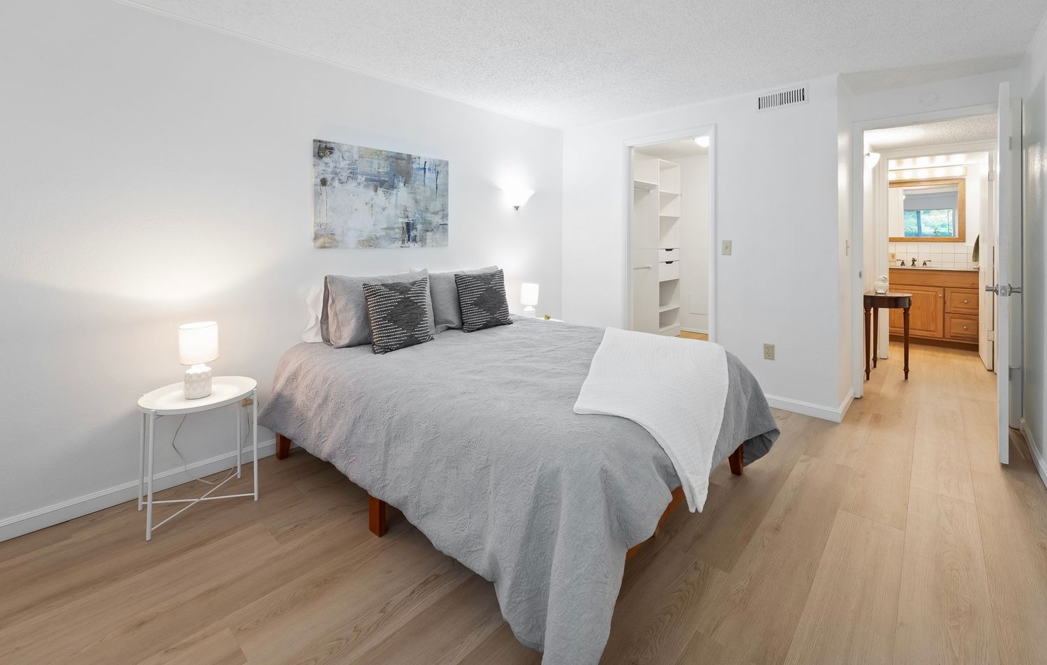Modern bedroom with gray bedding, light wood floors, white walls, and an en-suite bathroom visible in background.
