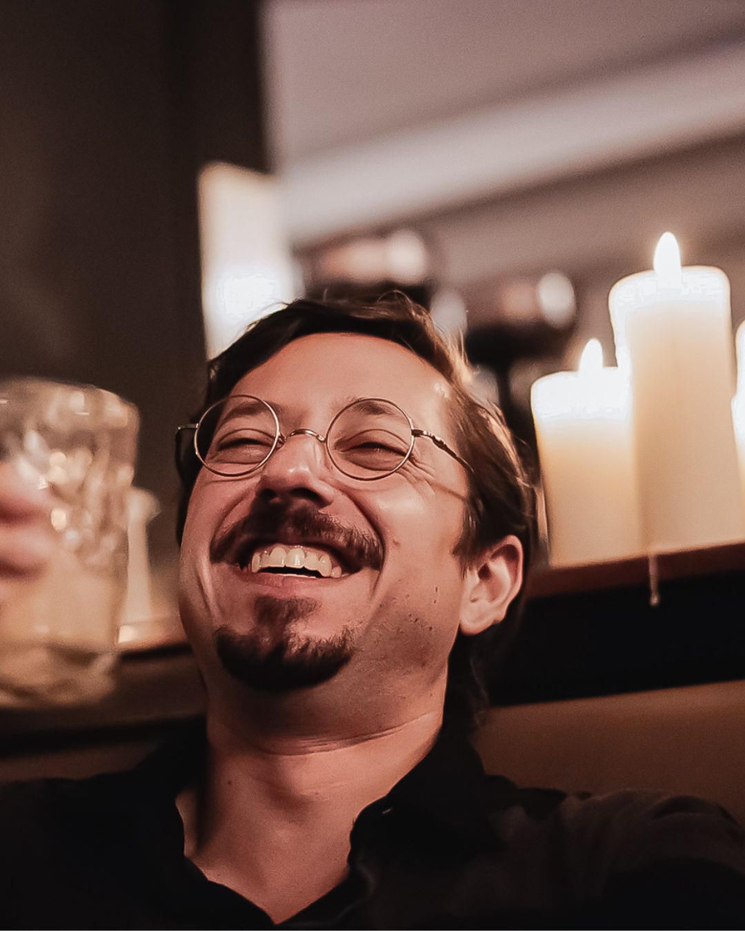 Cheerful bearded man with round glasses laughs near glowing white candles in warm candlelit setting.