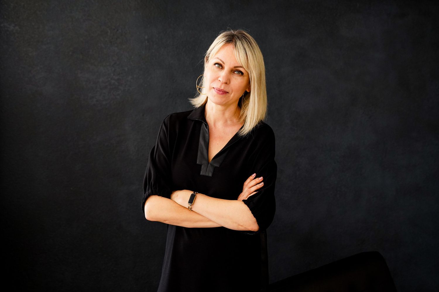 A photo of a woman wearing a black dress and smiling at the camera she has her arms crossed and is against a black backdrop.