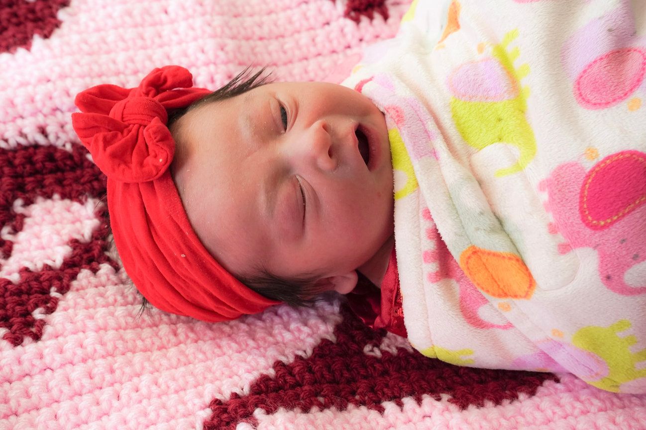 A photo of the baby laying on a pink blanket, the baby is wrapped in a blanket and wearing a red bowtie headband.
