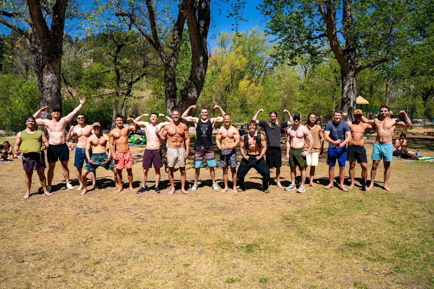 Large group of shirtless men flexing and posing outdoors in a sunny park with trees in the background.