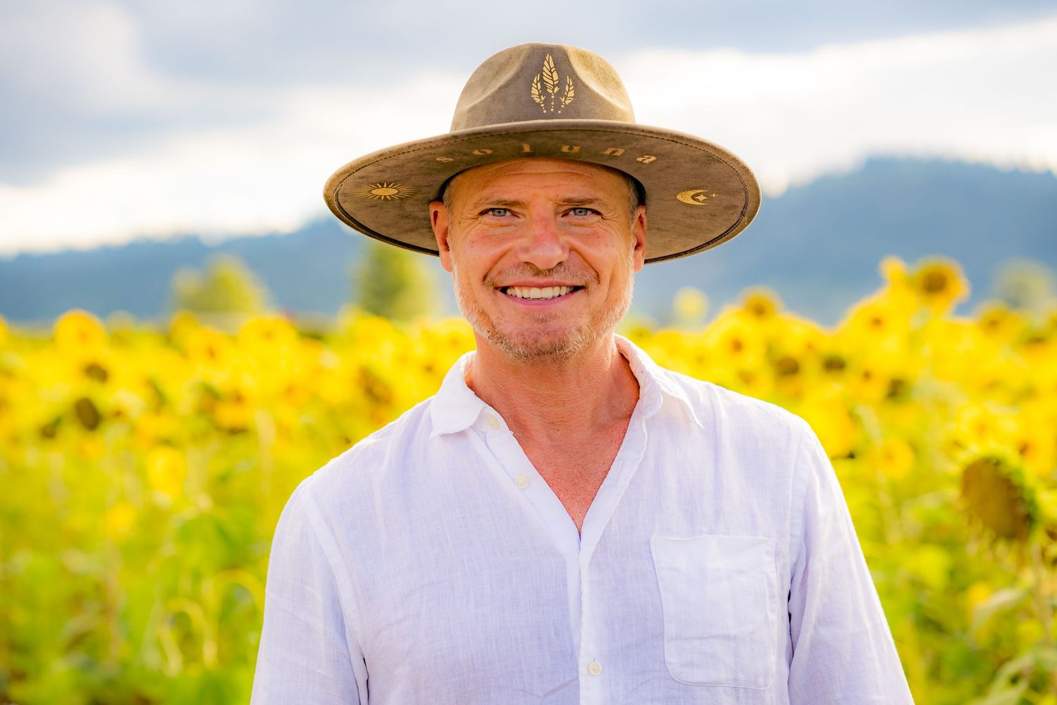 Smiling man in a wide-brim hat and white shirt standing in a vibrant sunflower field with mountains in the background.