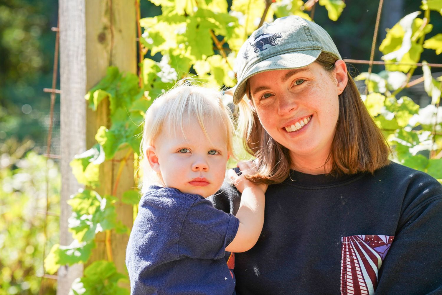 A smiling woman in a cap holds a blonde toddler outdoors near green grapevines on a sunny day.