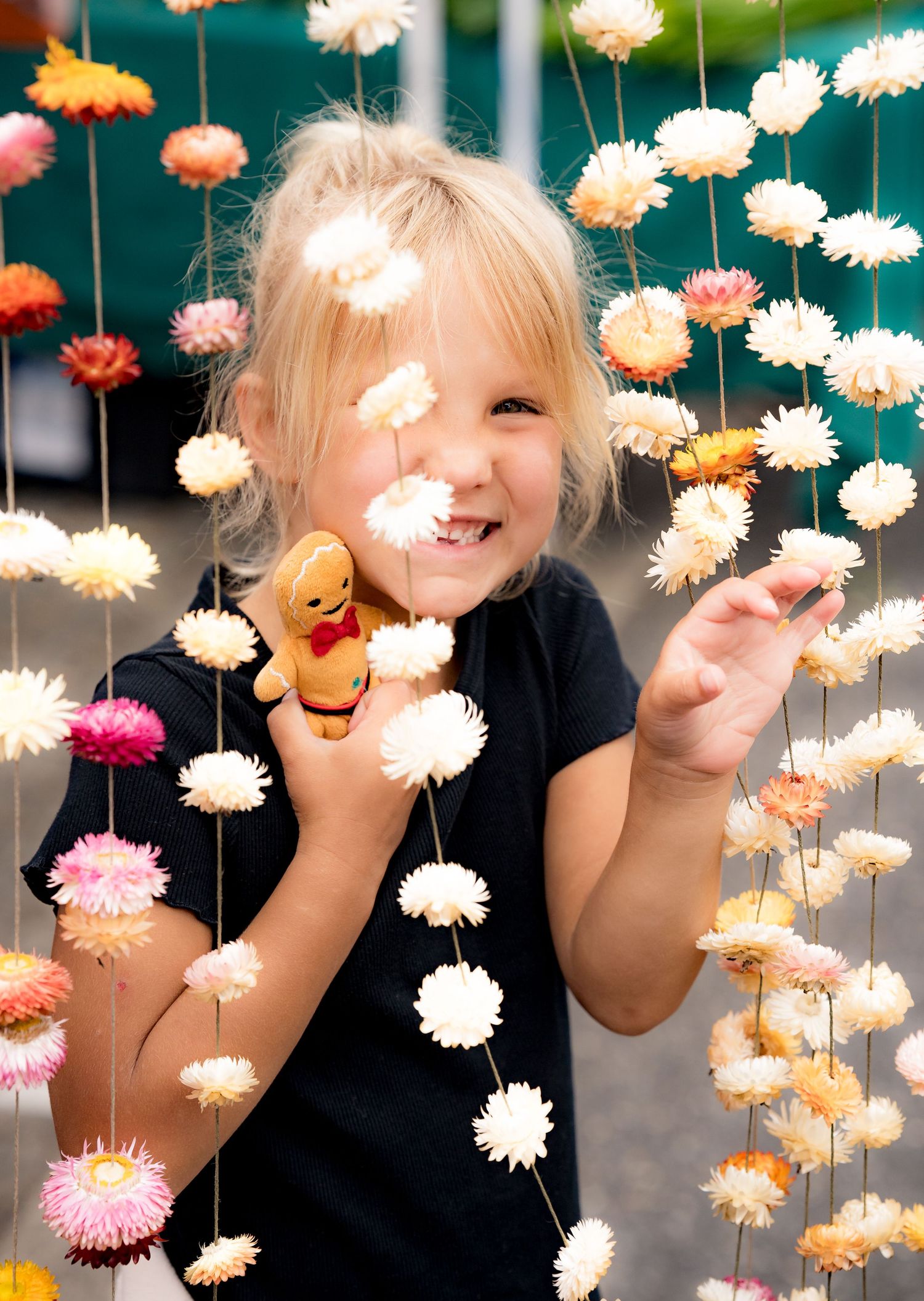 Smiling blonde girl holding a toy gingerbread man among hanging floral garlands with pink and white flowers.