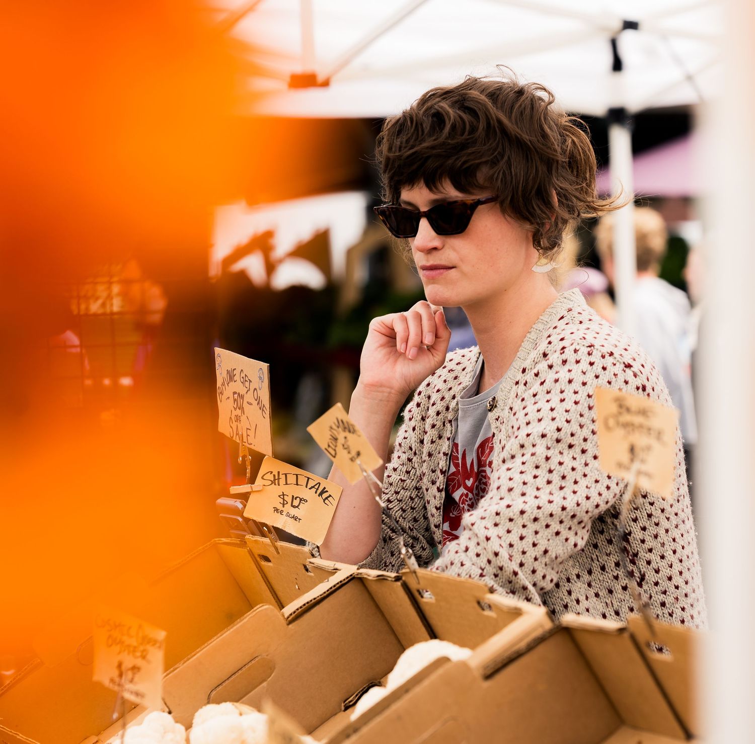 Woman in sunglasses and polka dot blouse browsing cardboard boxes at an outdoor farmers market.