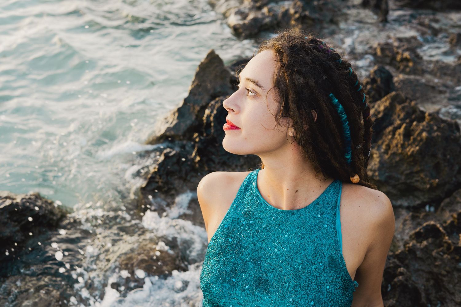 Person in a teal dress poses thoughtfully by rocky ocean coastline with crashing waves at sunset.