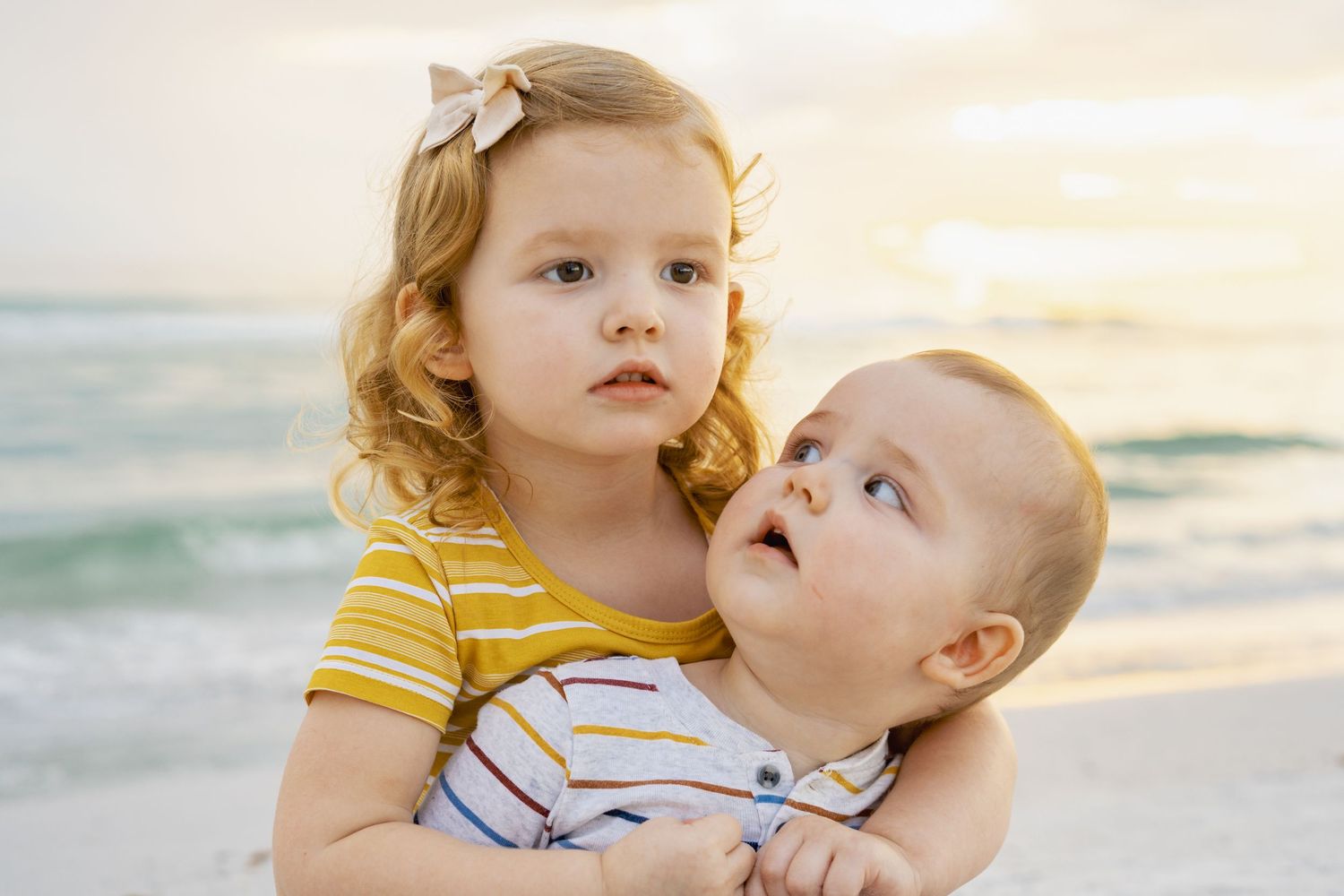 Children in coordinated yellow and white outfits embrace on a sunny beach at sunset.