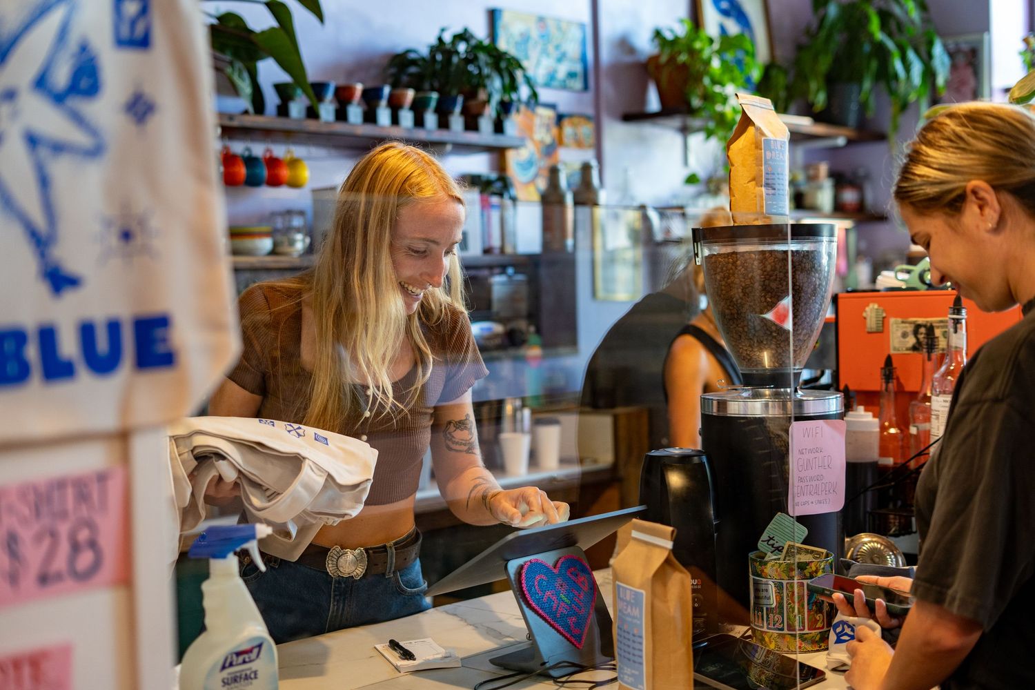 Barista with blonde hair serves customer at a cozy coffee shop counter with espresso equipment.
