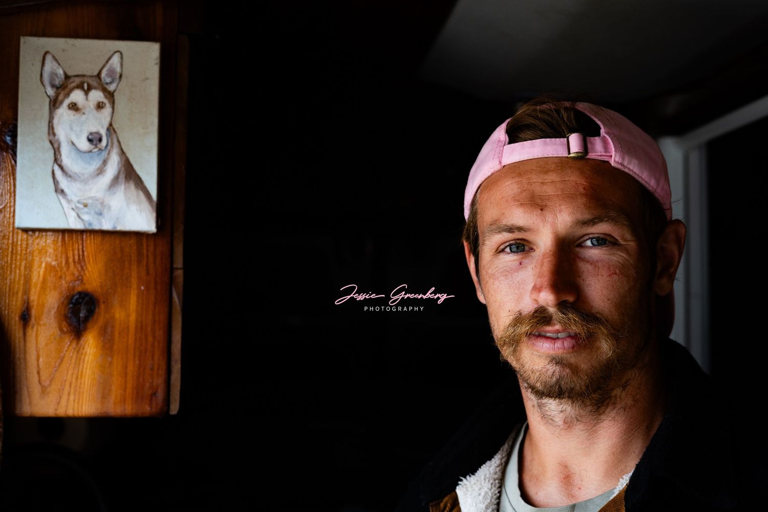Man with pink cap and mustache poses indoors beside a framed husky dog portrait on a wooden wall.
