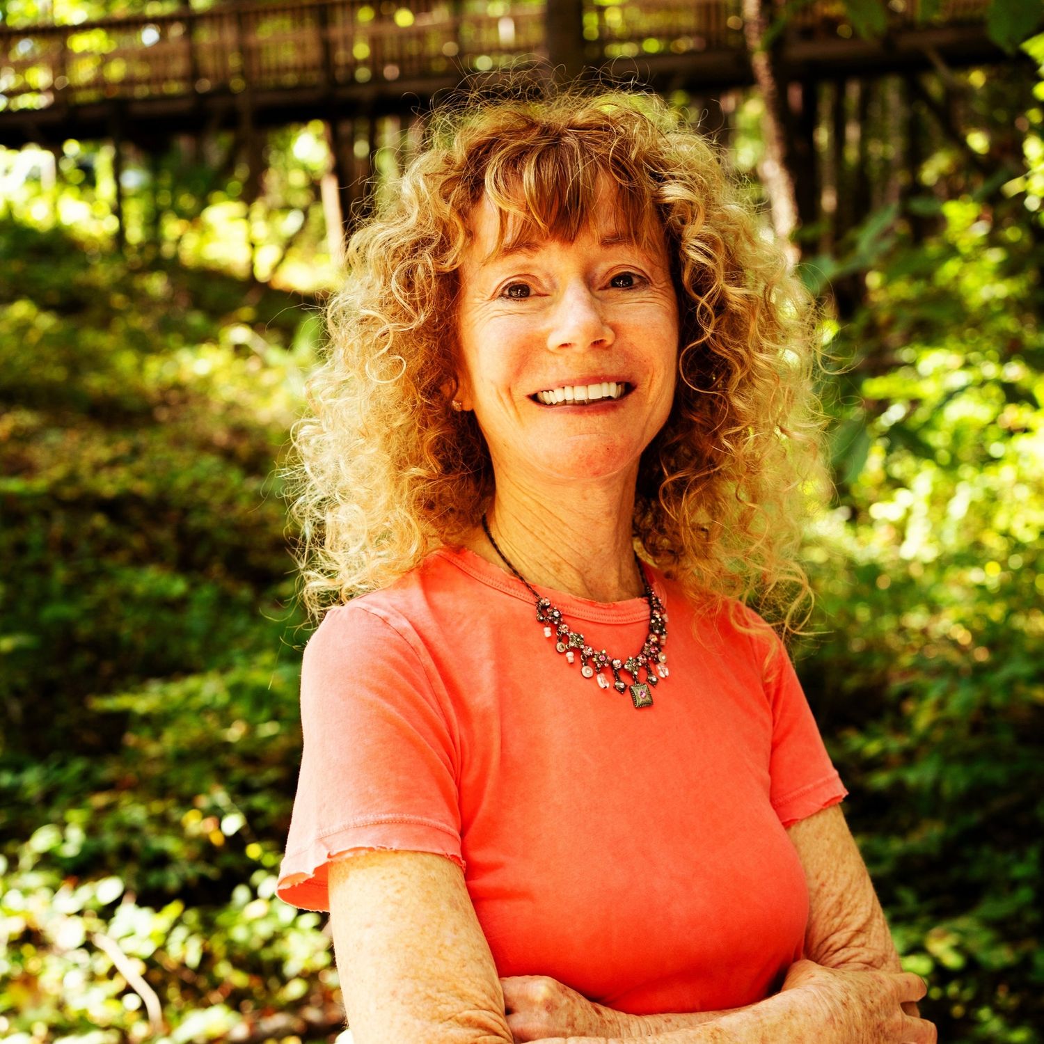 A picture of a woman with red curly hair smiling at the camera, she is wearing an orange shirt and a necklace and has greenery behind her.