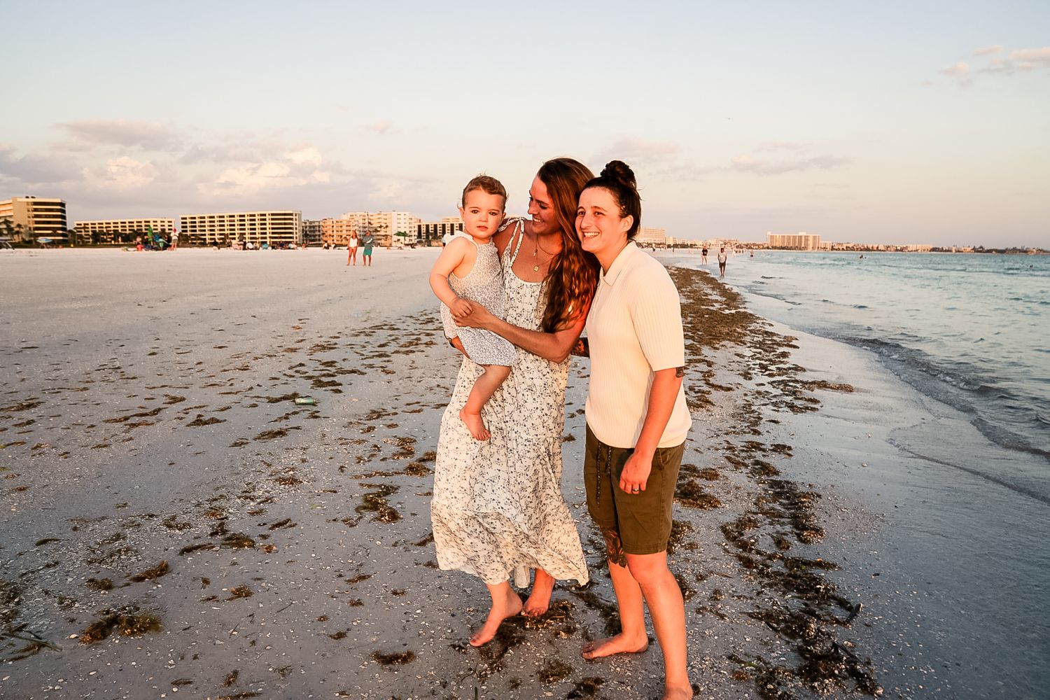 A photo of wives with their baby girl by the water on the beach with rainbow sky behind them.