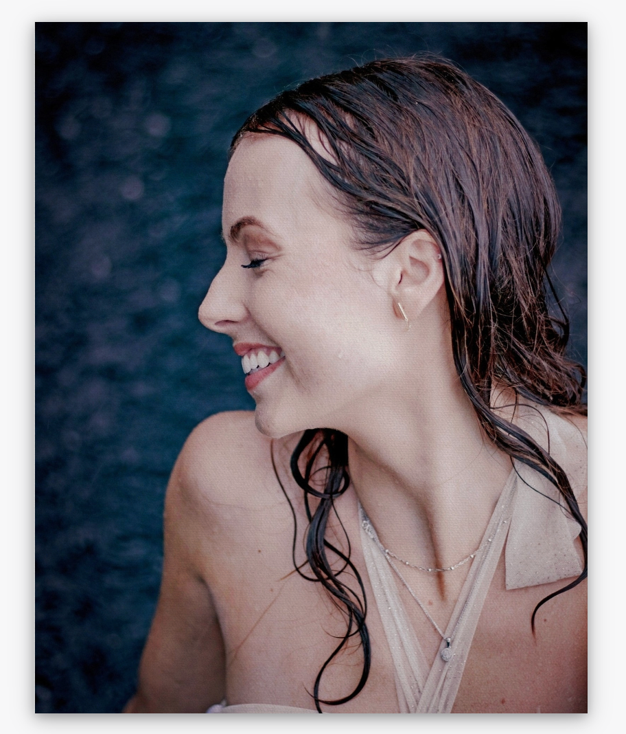 A photo of the girl smiling and looking at the water, she is wet and wearing a white dress.