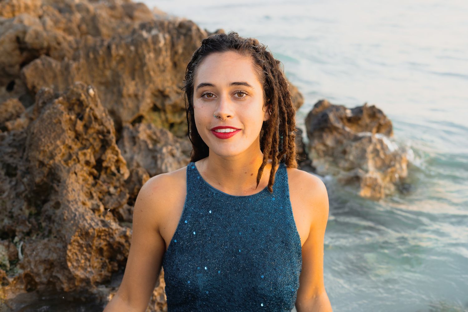 Young woman with dreadlocks in a blue sequin top posing by rocky ocean shore at golden hour.