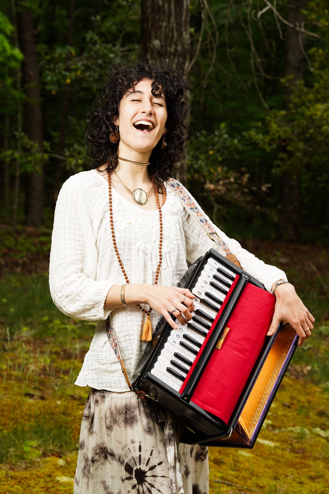 A joyful curly-haired woman plays a red accordion outdoors in a lush green forest.