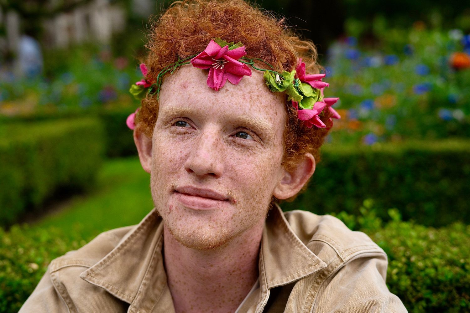 Young man with red curly hair and freckles wearing a pink flower crown in a garden.