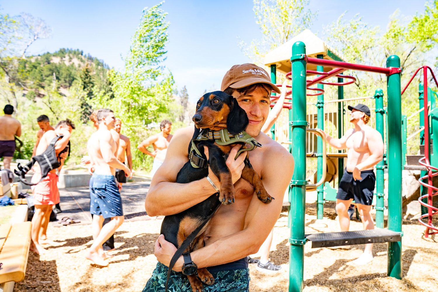 Shirtless man holding a dachshund dog at an outdoor playground on a sunny day.