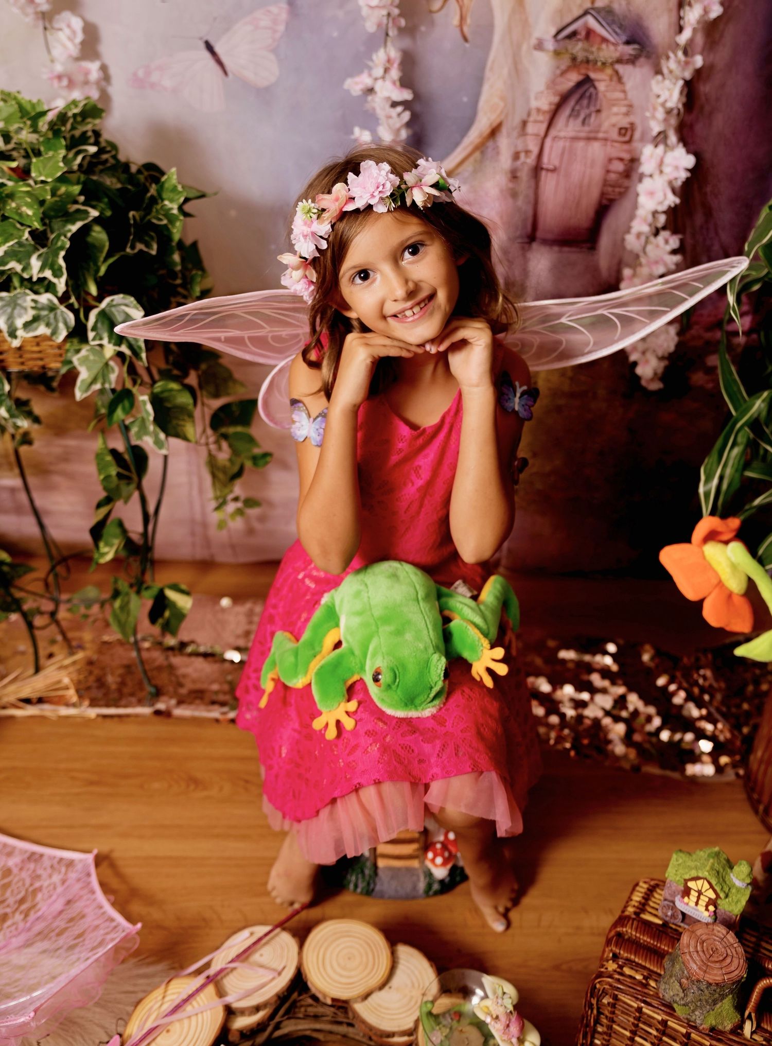 Young girl in pink fairy costume with wings, flower crown, holding green frog plush in enchanted forest setting.