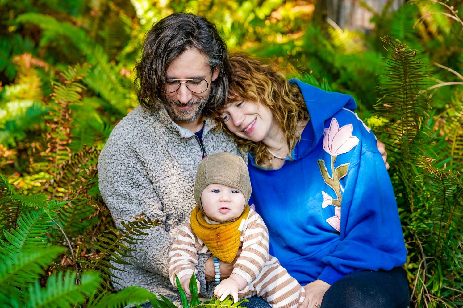 A couple with curly hair sits among lush ferns holding their baby dressed in a mustard outfit in a forest setting.