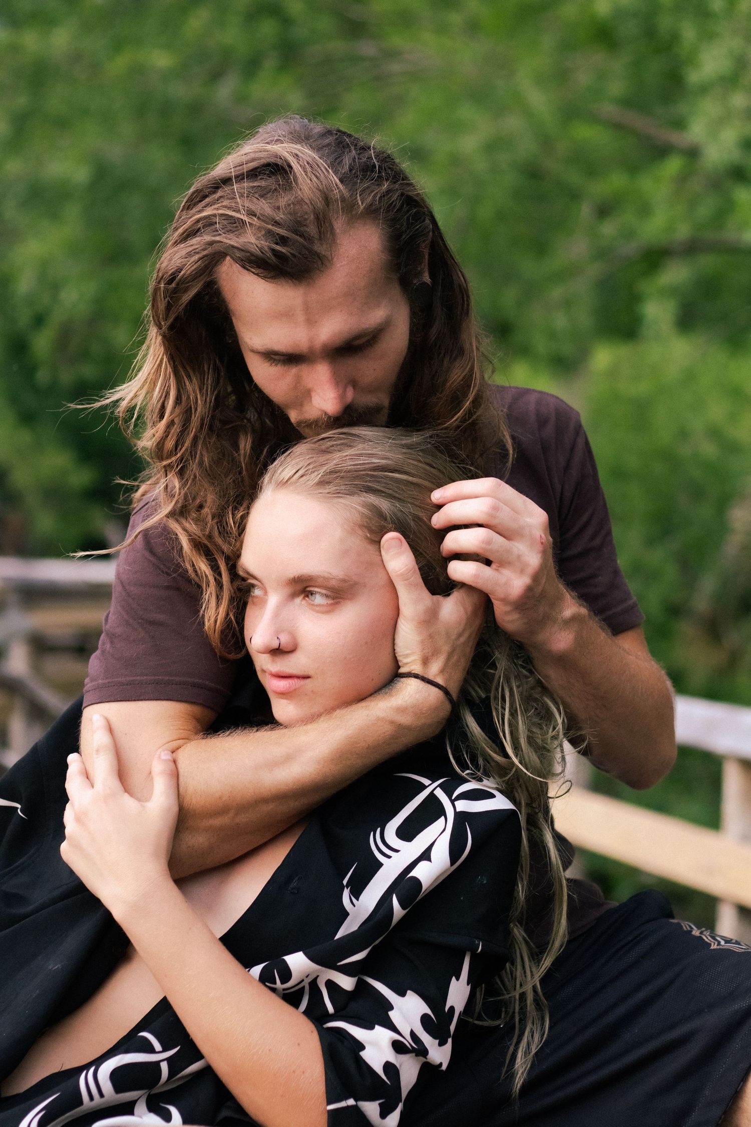 A man with long hair tenderly embraces a blonde woman outdoors, kissing her head against a lush green background.