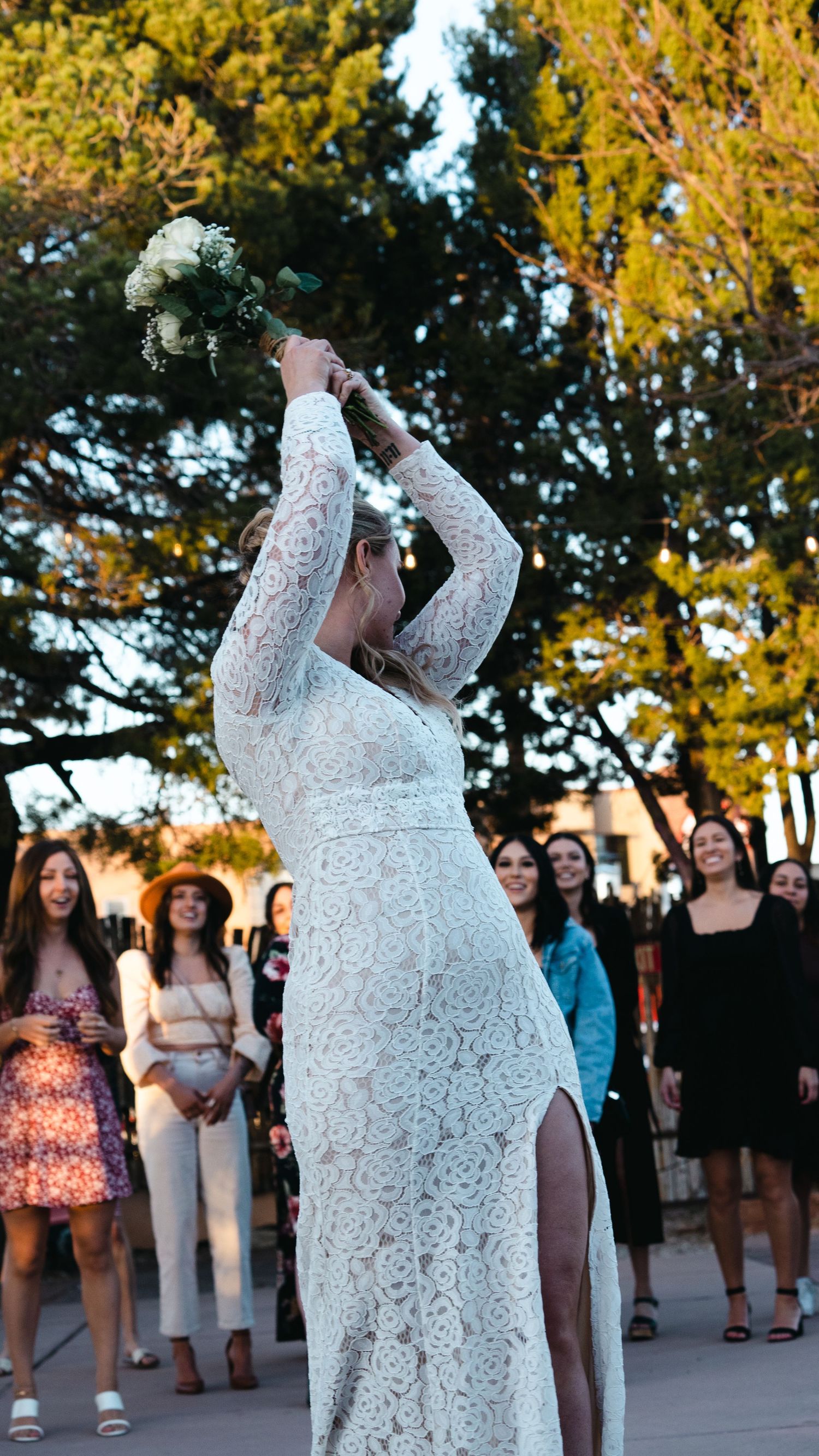Bride in white lace gown tossing bouquet to guests at outdoor wedding ceremony.