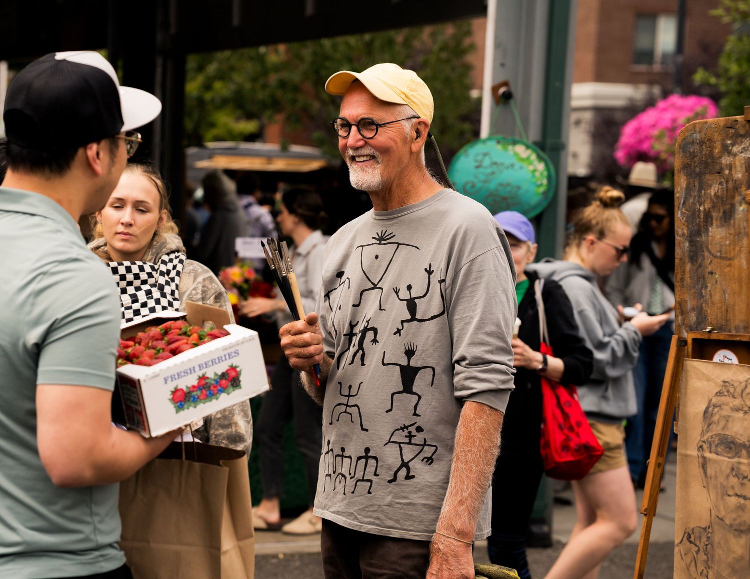 Older man with glasses and yellow cap smiling at outdoor farmers market, holding paintbrush near an easel.