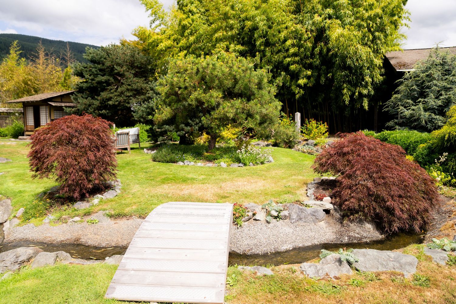 A serene Japanese garden with a wooden bridge, red maple trees, lush greenery, and a traditional structure in the background.