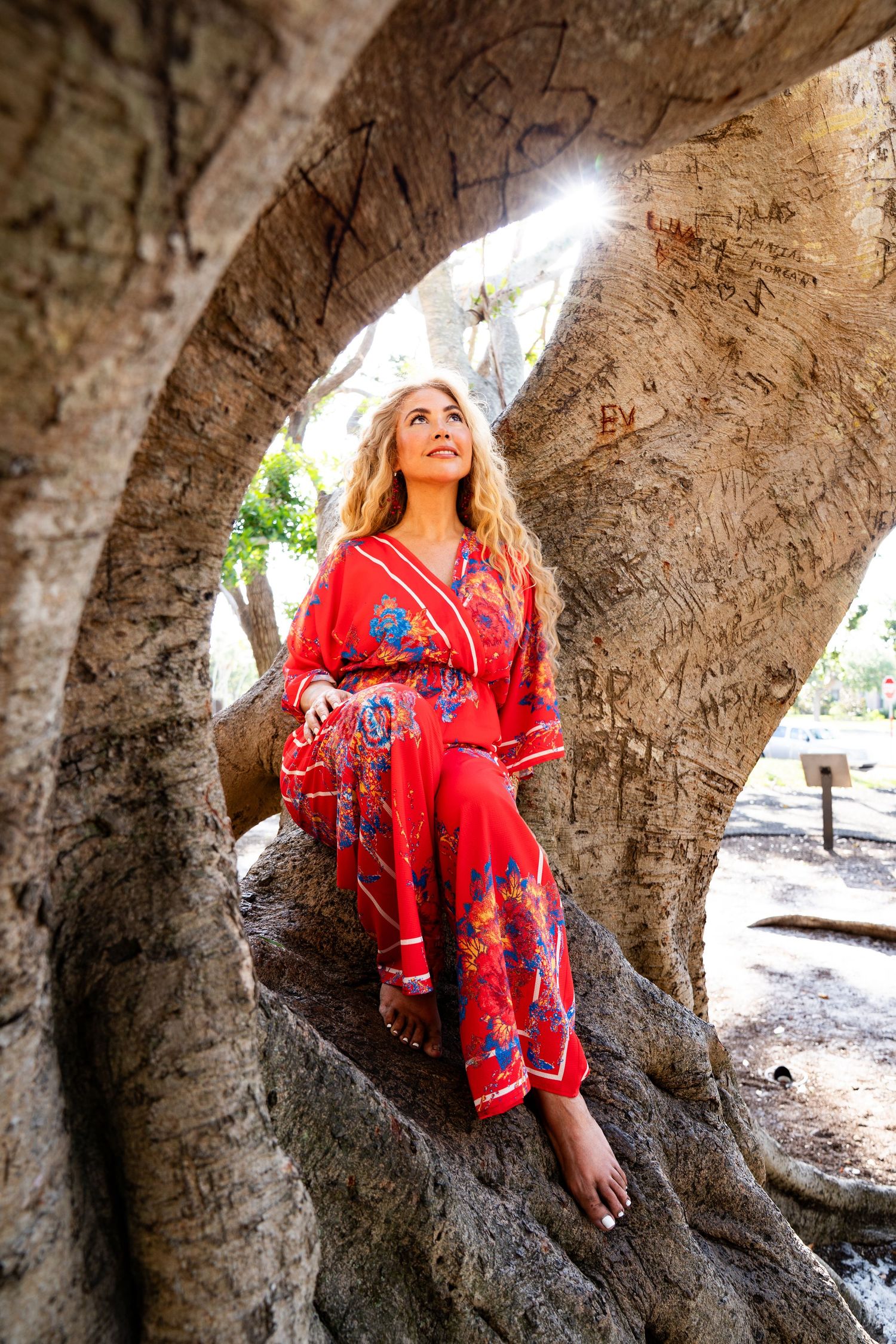 A photo of a woman wearing a red outfit sitting on a tree with the sunlight peaking through.