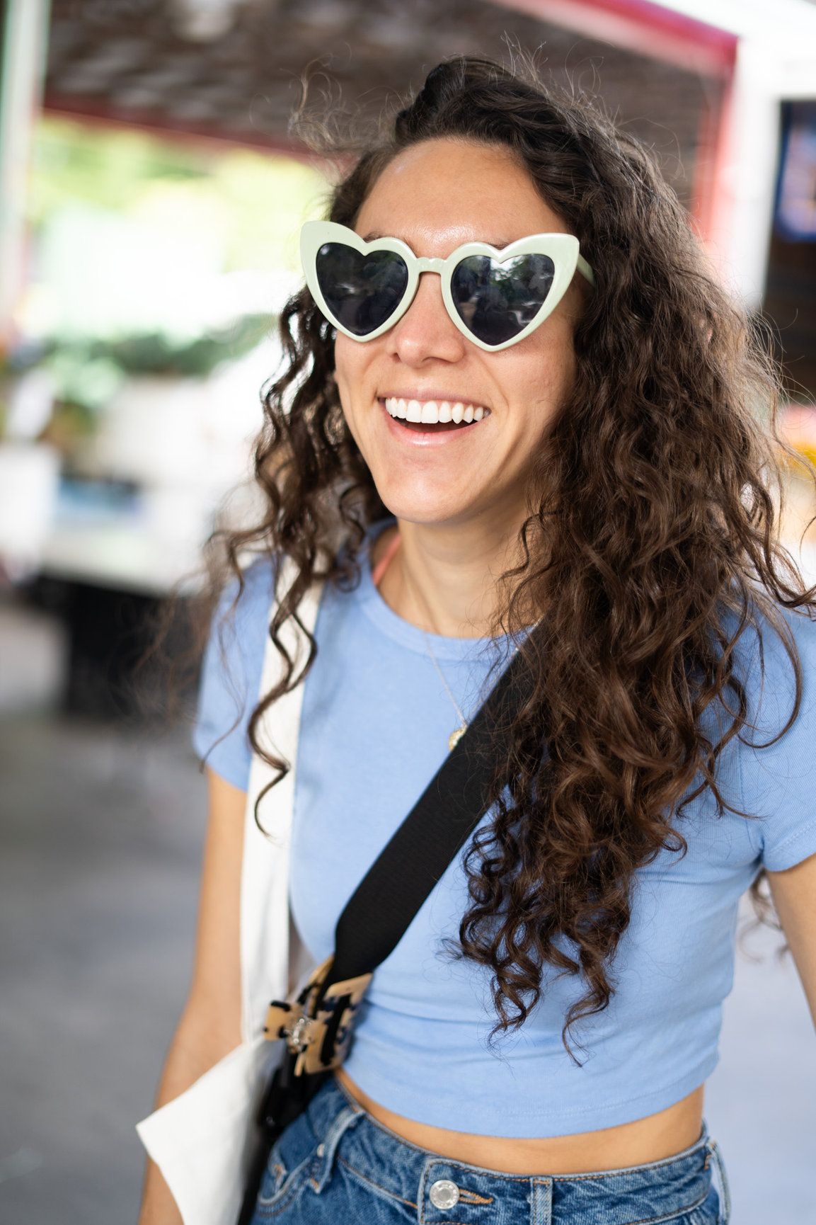 A photo of a woman with brown curly hair wearing heart white sunglasses laughing, she is wearing a light blue top and is having fun
