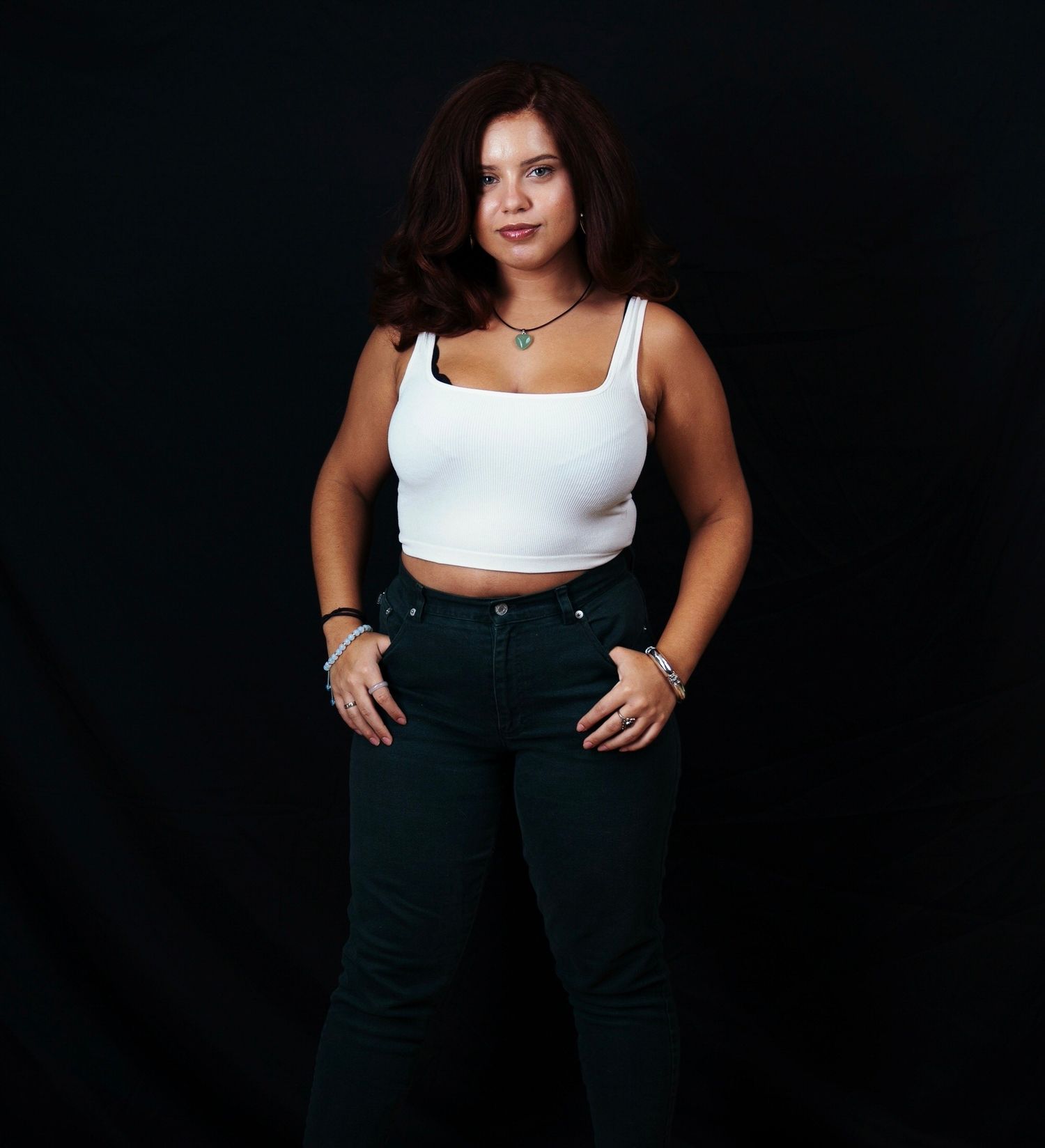 Young woman in white crop top and dark jeans posing confidently against a black background.