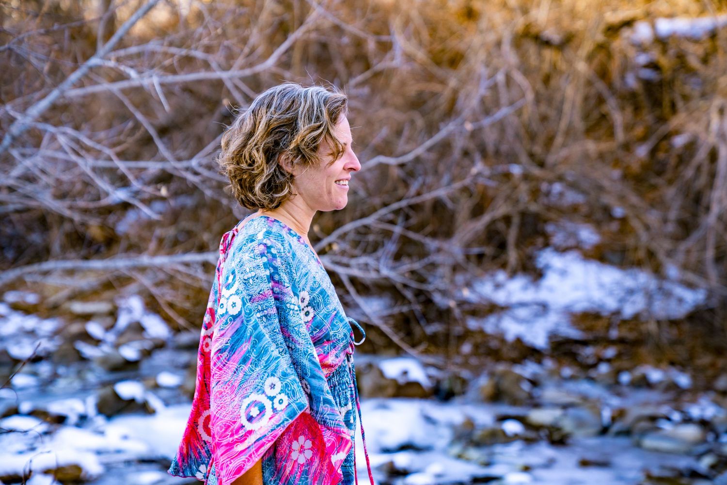 A person wearing a colorful patterned coat walks along a snowy creek bank in winter.