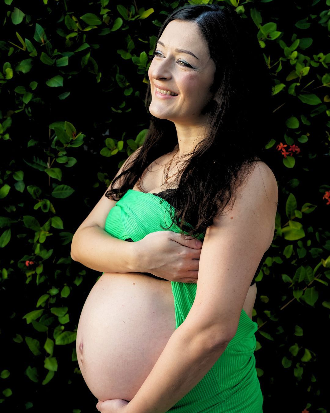Smiling pregnant woman in green dress cradling her baby bump outdoors against lush green foliage backdrop.