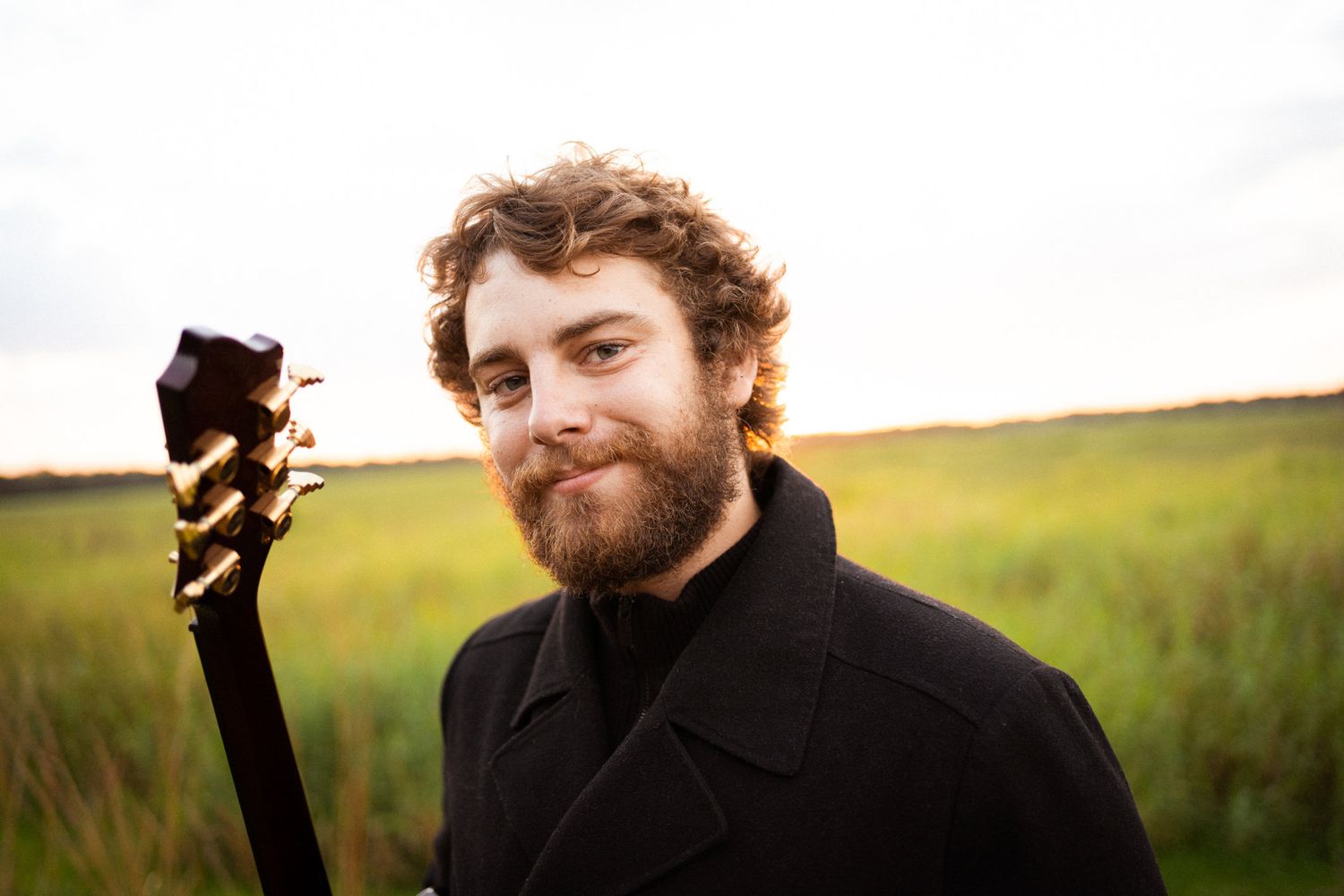 A bearded man wearing black, holding his guitar and looking at the camera.