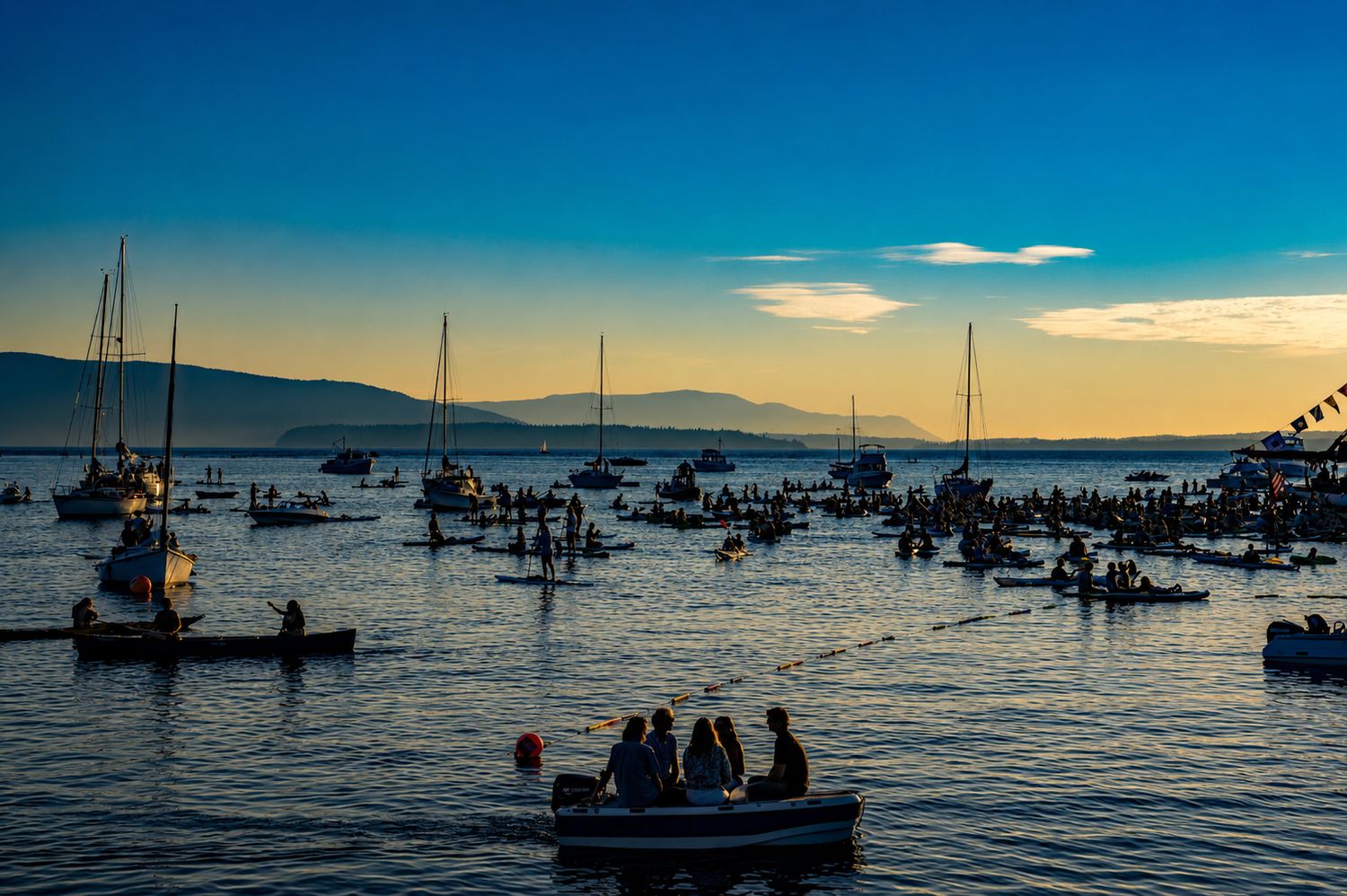Boats and sailboats fill a calm bay at sunset with mountains in the background and people gathered on the water.