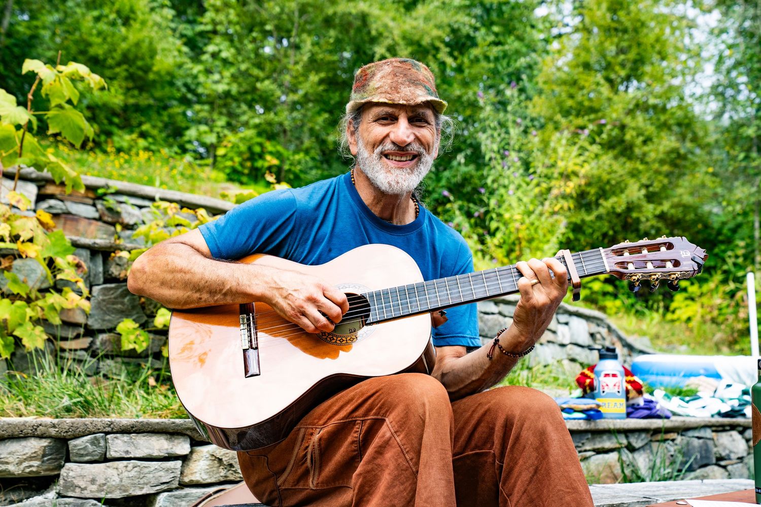Smiling older man with beard playing acoustic guitar outdoors near stone wall surrounded by green trees.