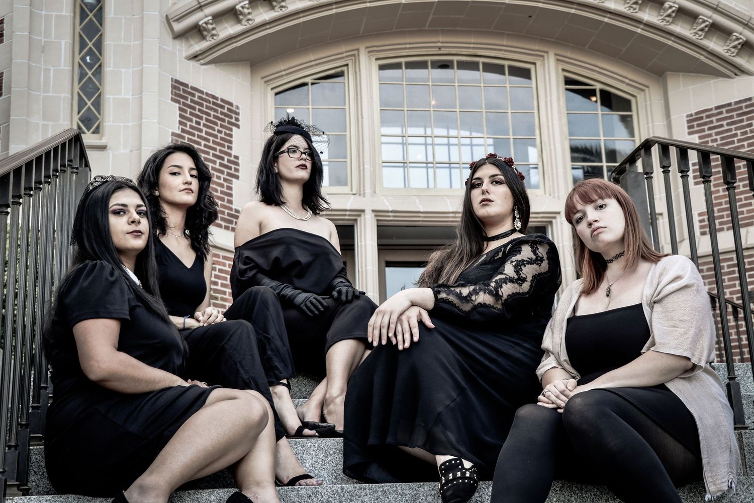 Five women in black dresses pose on stone steps outside a brick building for a stylish group photo.