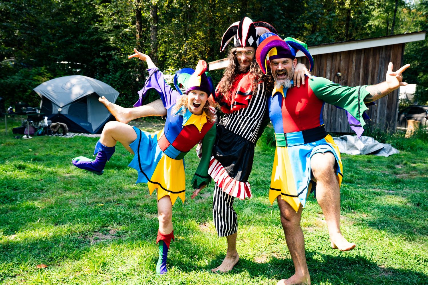 Three people in colorful jester costumes pose playfully on green grass at an outdoor festival campsite.