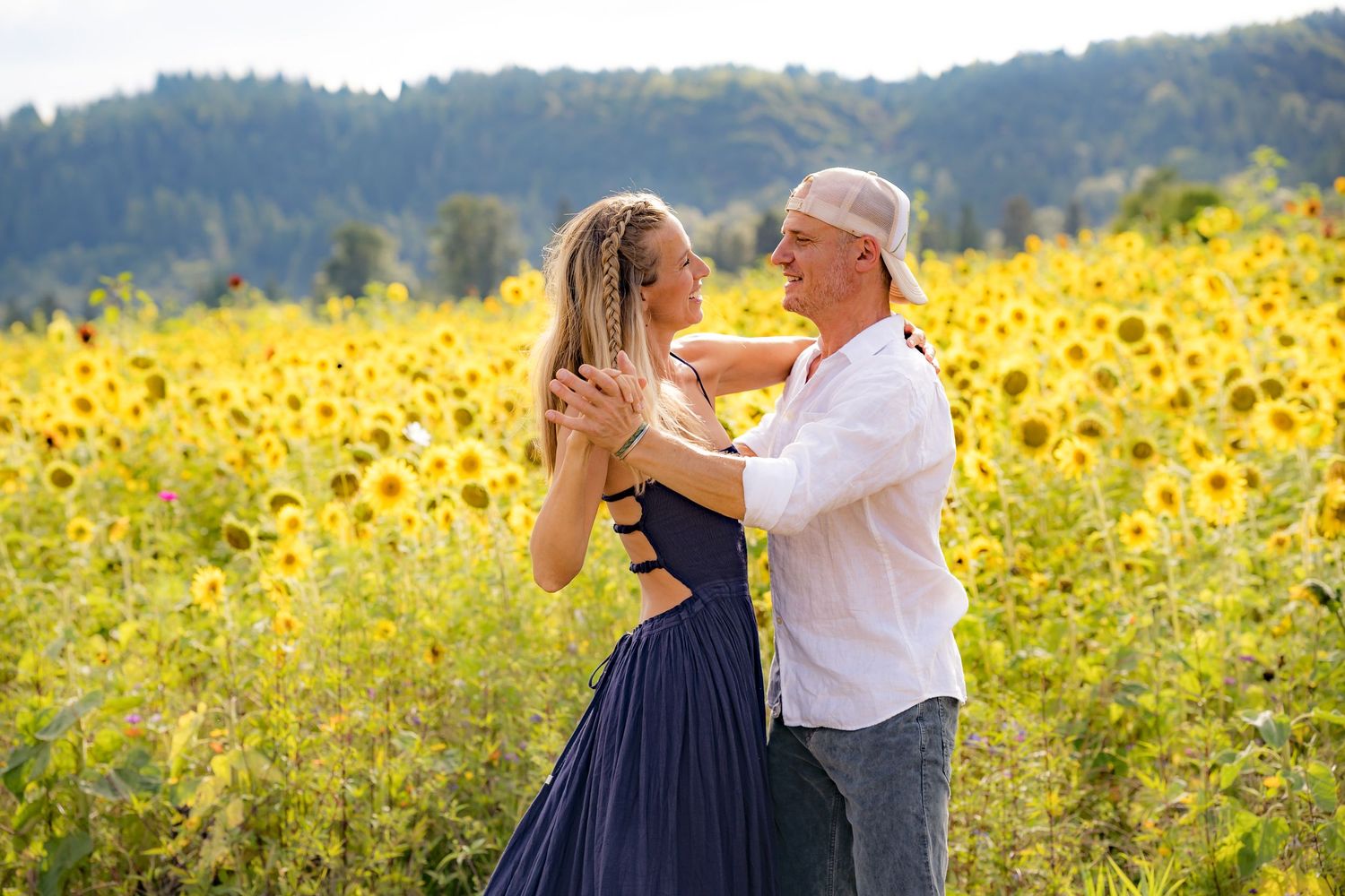 Couple dancing romantically in a vibrant sunflower field with lush green mountains in the background.