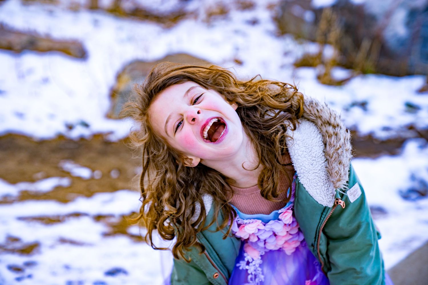 A person with curly hair wearing a green coat laughs joyfully in a snowy winter landscape.